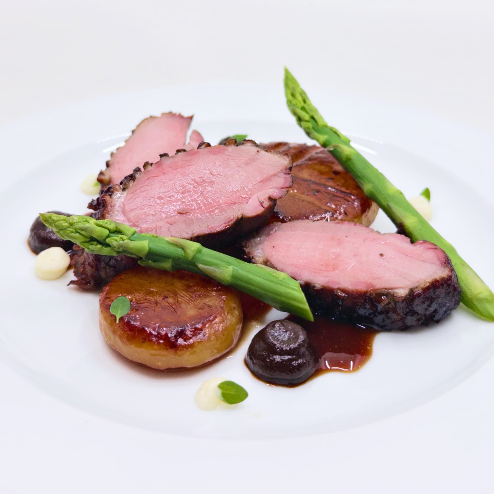 Three slices of pink meat with two asparagus spears arranged at odds, opposed to glossy potatos pucks with dollops of white and brown gel and a glossy sauce with microgreens on top