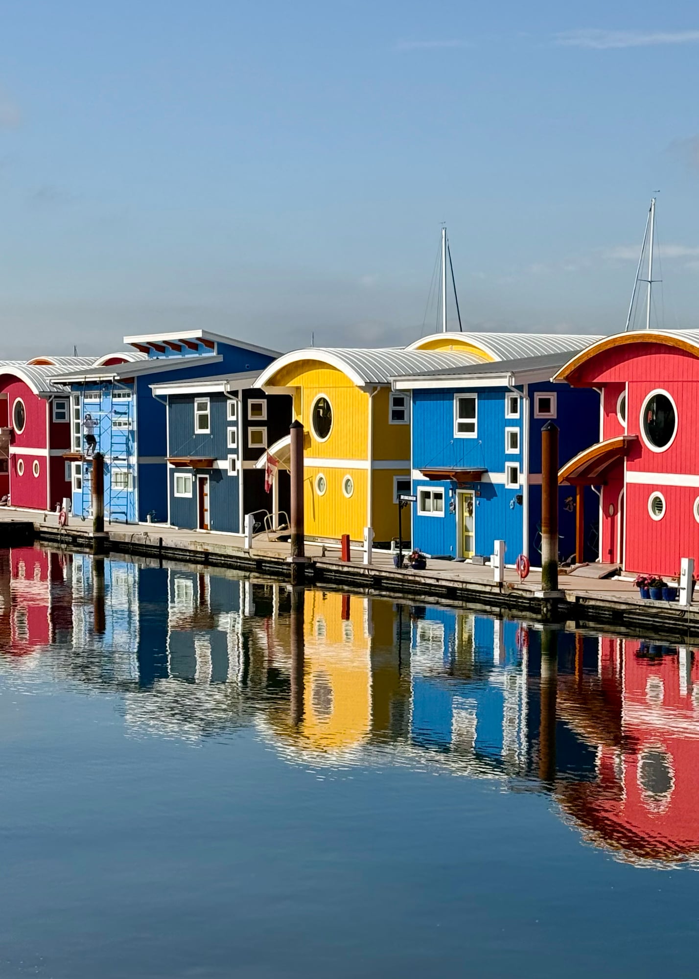 Row of bright red, yellow, and blue two-storey hosues running diagonally toward the viewer, reflected in the ocean they sit on broken only by the wooden walkway leading to each.