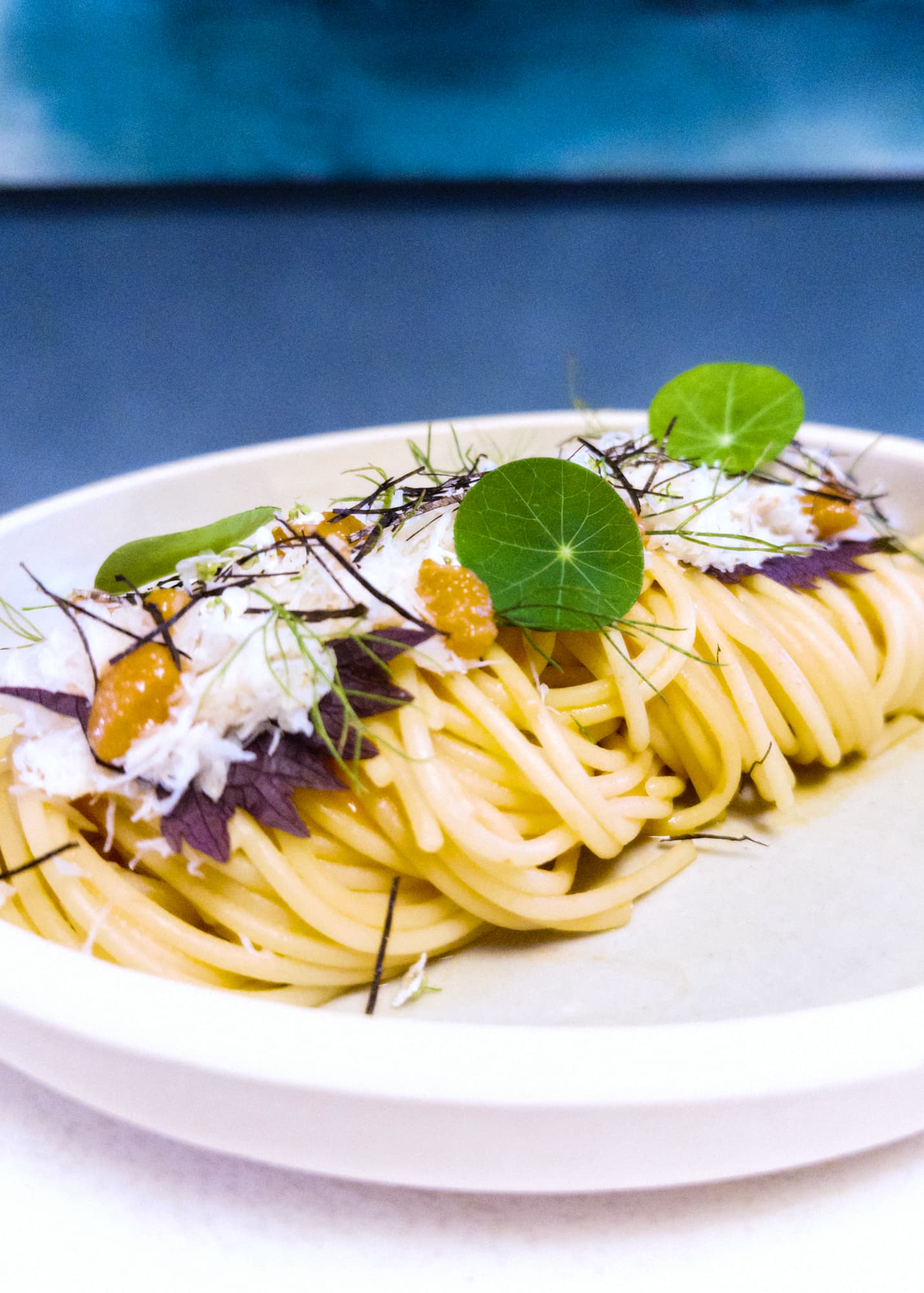 Twirled spaghetti topped with white meat, jagged purple leaves, green leaves and pink dollops in a rimmed plate against a blue background.