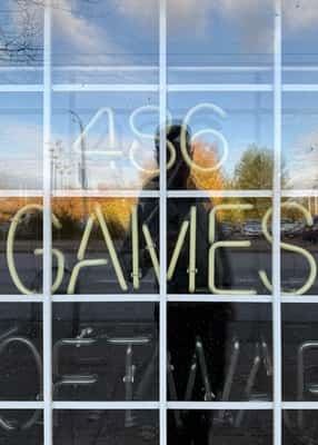 Neon sign reading 486 GAMES seen through a bar-lined window, reflection of the photographer