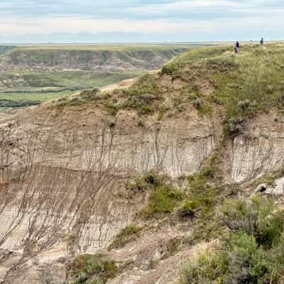 Canyon rimmed with reddish-tinged ragged layered clay hills topped in low green scrub under a cloudy sky. Foreground is a large hill with tiny people on top to indicate scale.