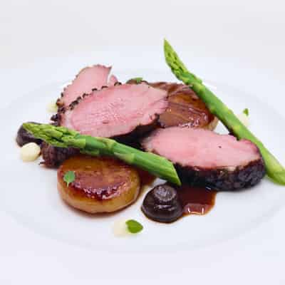 Three slices of pink meat with two asparagus spears arranged at odds, opposed to glossy potatos pucks with dollops of white and brown gel and a glossy sauce with microgreens on top