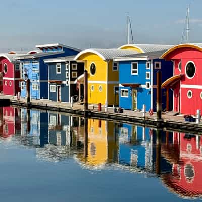 Row of bright red, yellow, and blue two-storey hosues running diagonally toward the viewer, reflected in the ocean they sit on broken only by the wooden walkway leading to each.