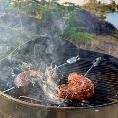 Two skewered curved steaks on a smokey grill with trees, rocks, and ocean in the background
