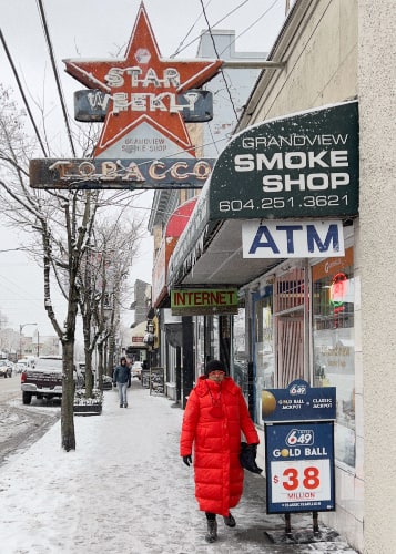 Snowy sidewalk being used by a woman in a puffy red coat under a faded neon sign saying 'Star Weekly / Tobacco'