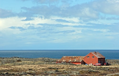 Red barn-type building on barren rocks covered in low scrub with a very blue ocean and sky behind