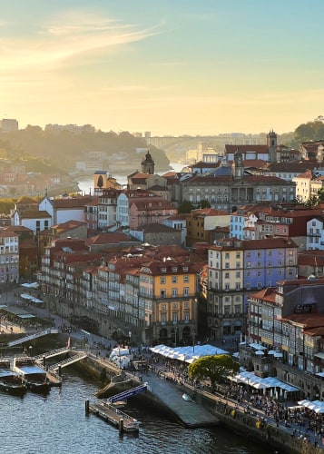 Hazy blue/yellow sky above classic Portuguese buildings on a riverbank crowded with people