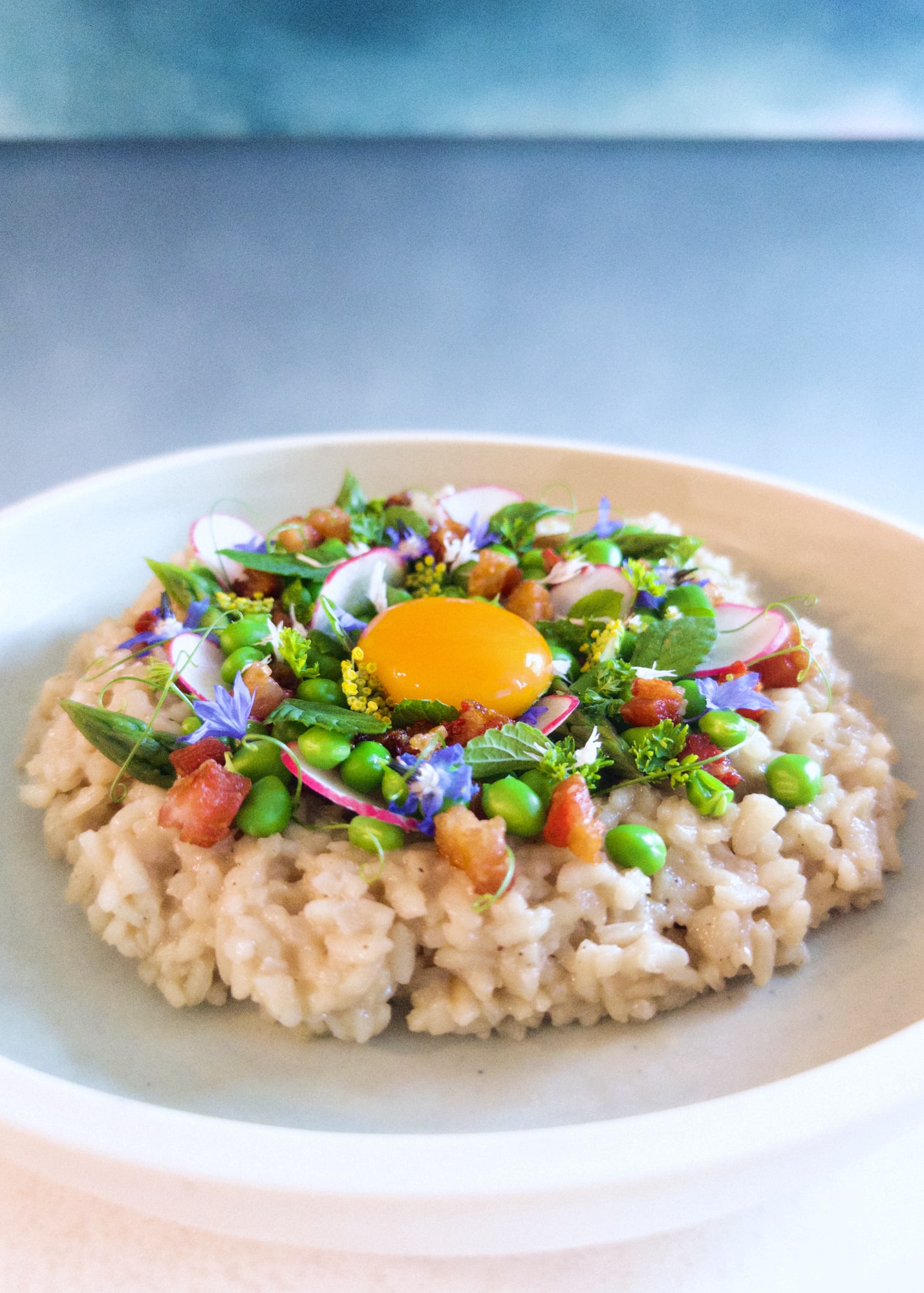 Rice arranged in circular shape topped with ornate green leaves and vegetables, pink radishes and guanciale chunks, purple flowers, and a yellow egg yolk in the center