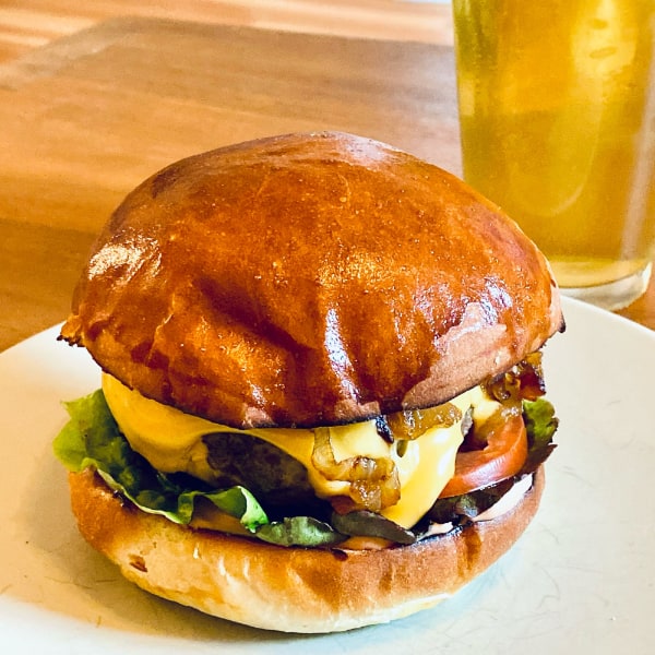 Cheeseburger with lettuce, tomato, and caramelized onion on a plate with a glass of pale yellow beer in the background