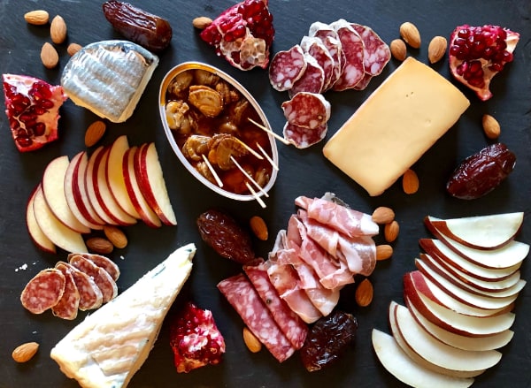 Grey slate board topped with an arranged assortment of cured meat slices, cheeses, fruit, nuts, and a tin of conservas