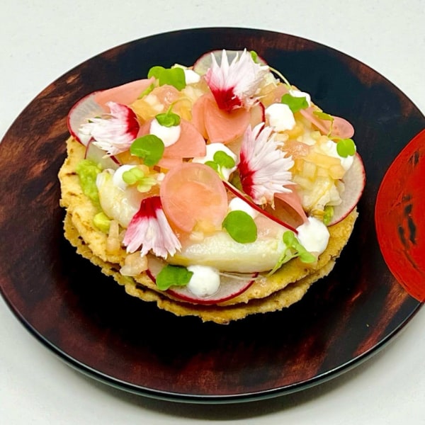 Double-stacked tostada topped with crab, pink pickled and raw red radish, white sauce, leaves and flowers on a wooden plate with a red painted semi-circle on one side
