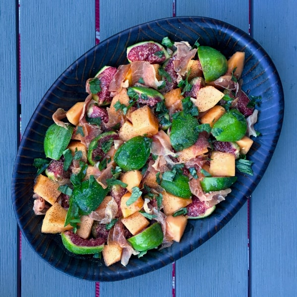 Oval plate on a blue table filled with mixed fruits, meat slices, and herbs