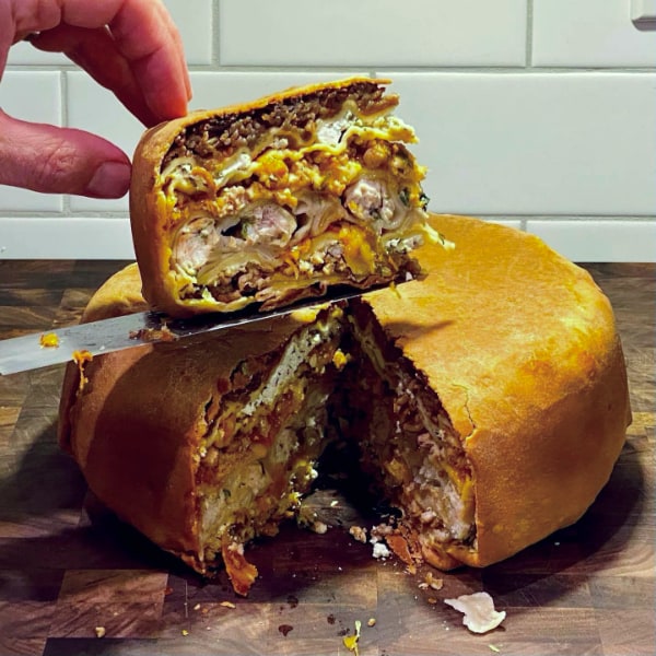 Hands balancing a thick slice of complex multi-layered, dough-wrapped food on a knife blade above the rest of the wheel it was cut from that sits on a wooden cutting board