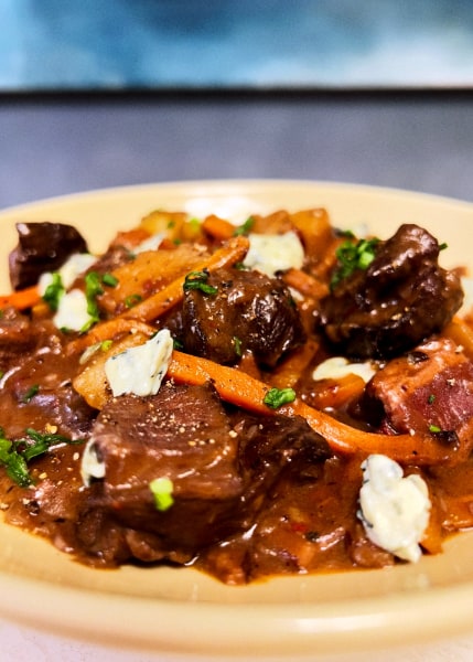 Glistening browned meat with carrots and cheese chunks in a rich brown sauce housed in a yellow bowl against a blue background