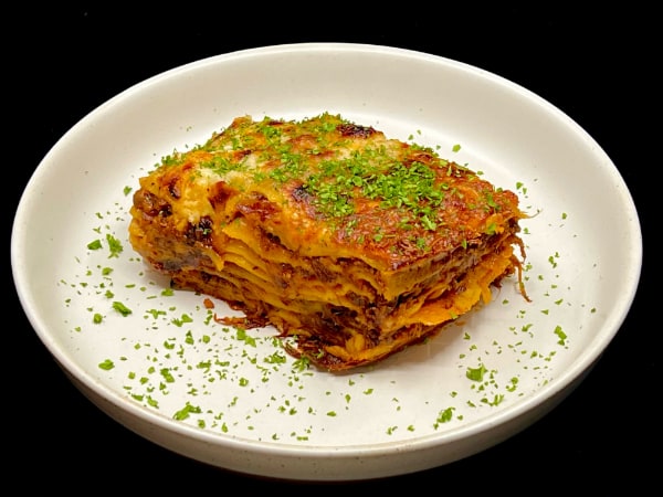 8-layer lasagna sprinkled with chopped parsley on a white rimmed plate against a black background