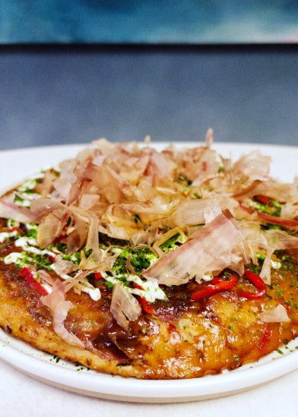Crispy golden pancake topped with red ginger slices, green onion, mayo drizzle and pink shaved bonito against a blue background