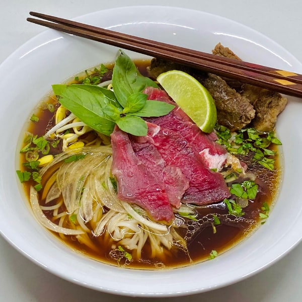 Pink slices of rare beef and brown well-done beef on noodles, herbs, and onion slices in a rich brown broth. In a white bowl with ornate brown chopsticks against a high-key white background.