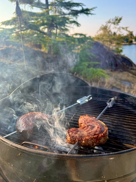 Smoking grill with two crescents of skewered steak mid-cook on a rocky tree-lined bluff in front of the ocean