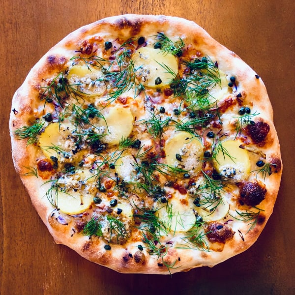 White pizza on blistered dough with potato slices, cheese, capers, and dill garnishing on a wooden surface