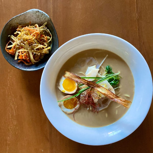 Small ornate bowl with mixed salad of long strips of various vegetables top right, opposed to large white bowl of cloudy ramen topped with egg halves, greens, lotus slices, and thin crispy brown strips.