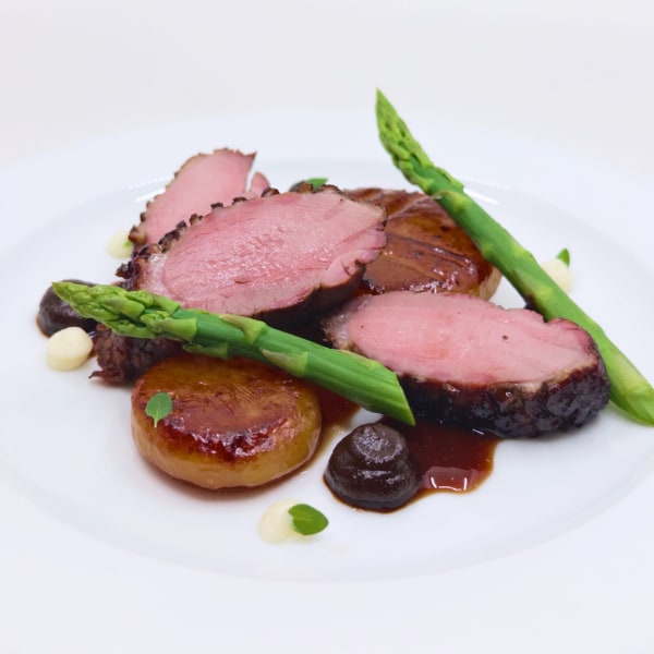 Thick slices of pink meat arranged with glistening browned discs, asparagus spears, and dollops of brown and white sauces on a white plate against a high-key white background