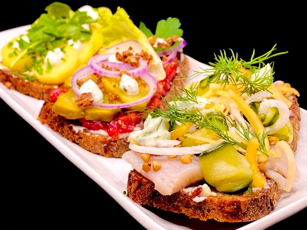 Trio of dark brown rye bread with unique toppings including pale fish slices, various pickled vegetables, pink tartare, greens, and onion slices