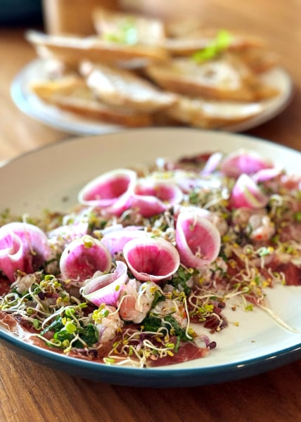 Curled pink watermelon radish, prawn chunks and sprouted greens on pink sliced meat in a shallow white plate, blurred bread slices in the background