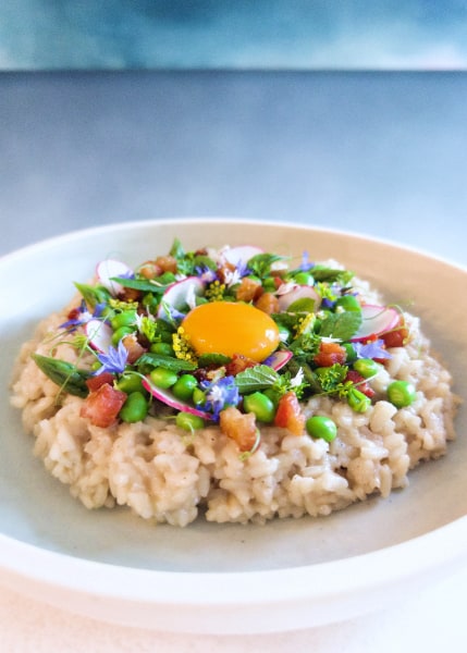 Disc of rice in a plate against a blue background with an ornate topping of green peas and herbs, asparagus tips, radish slices, crispy pink cubes, flowers, and a central egg yolk