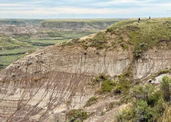 A few tiny figures on a grassy bluff top right overlooking layered strata descending down into a valley far below under a cloudy sky