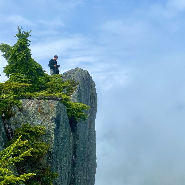 Solitary figure atop a sheer cliff face on the left side of the frame with nothing but clouds filling the rest of the view