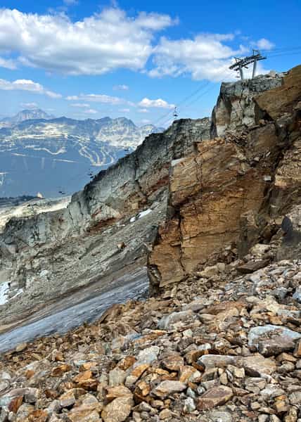 Orange-tinged boulder field in front of steep rocky cliffs with a chairlift running diagonally out of frame along the ridge, blue jagged mountains with evident ski trail cuts through the forest behind.