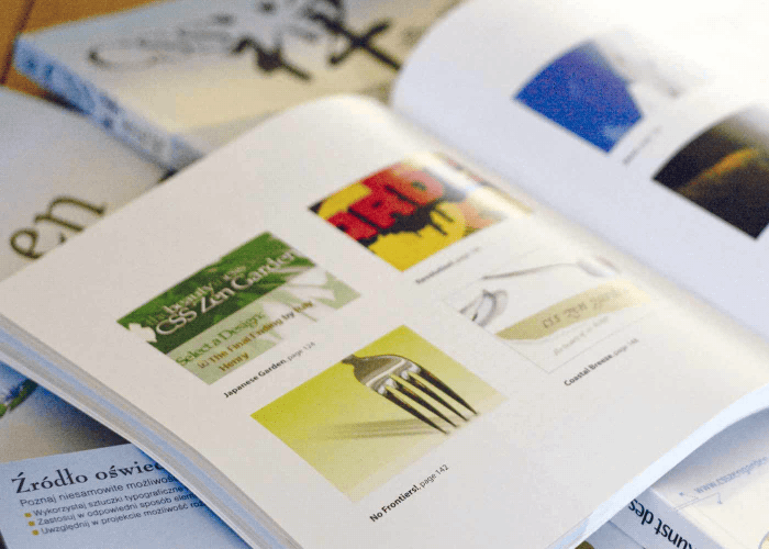 Book open to a page showing multiple designs sitting on a pile of translated copies of the same book.